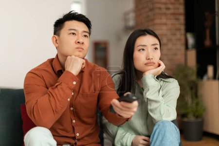 Foto de Discontented asian couple watching television, pointing remote controller and switching TV channels, sitting on sofa at home. Boring movie, displeased viewers concept - Imagen libre de derechos