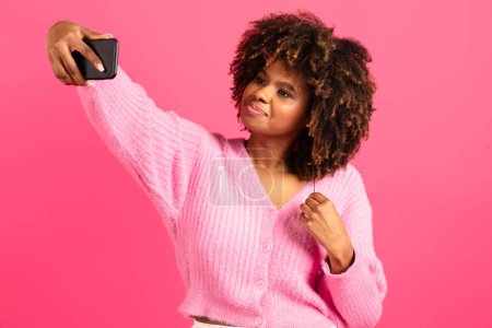 Téléchargez les photos : Cheerful young black curly lady student in casual takes selfie on smartphone, shooting video for blog and social media, isolated on pink background, studio. App for photo, ad and offer, beauty care - en image libre de droit