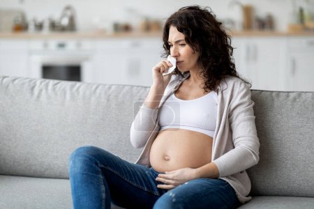 Téléchargez les photos : Sick Pregnant Woman Suffering Cold Or Flu While Sitting On Couch At Home, Young Expectant Female Feeling Unwell, Having Grippe Or Allergy, Touching Belly And Blowing Nose To Napkin, Copy Space - en image libre de droit