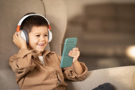 Téléchargez les photos : Happy little boy in wireless headphones watch video on smartphone, sits in armchair, calls on phone in room interior. Technology at home, app for communication, game, education for child and childhood - en image libre de droit