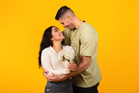 Téléchargez les photos : Cheerful young arab guy in casual hugs woman with flowers, enjoy romantic, isolated on yellow background, studio. Love, relationship, congratulations of holiday, Valentine day and anniversary, date - en image libre de droit