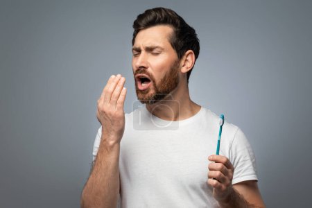 Téléchargez les photos : Bad breath. Handsome middle aged man checking his breath with his hand, blowing to it, standing over grey background. Bad smell from the mouth, toothache, having problems with teeth - en image libre de droit