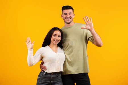 Téléchargez les photos : Cheerful young arab woman and guy in casual hugging, waving hands at camera, isolated on yellow background, studio. Love, relationship, video call, greeting, say hello, hi sooting video, ad and offer - en image libre de droit