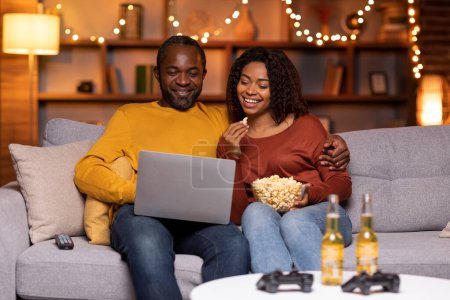 Foto de Happy cheerful married african american couple sitting on couch, using laptop at cozy winter evening, watching TV show, eating popcorn, drinking beer, resting at home together, free space - Imagen libre de derechos