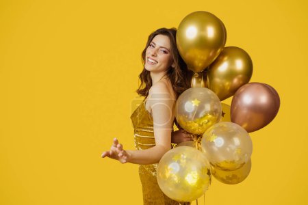 Photo for Festive concept. Charming lady in dress with bunch of air balloons having holiday party over yellow studio background, copy space. Cheerful woman having fun on her birthday celebration - Royalty Free Image