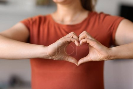 Téléchargez les photos : Unrecognizable Woman Making Heart Gesture With Hands Near Chest, Closeup Shot Of Young Female In T-Shirt Showing Love Sign At Camera, Expressing Kindness While Standing Indoors, Cropped - en image libre de droit