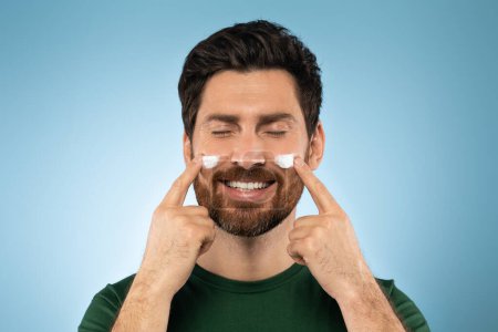 Photo for Happy bearded handsome man applying face cream on cheeks with closed eyes, posing over blue studio background. Good looking middel aged man using beauty products for his face - Royalty Free Image