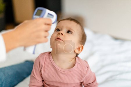 Foto de Healthcare for infant child. Mother measuring temperature of her baby girl with infrared thermometer, kid looking at moms hand, closeup - Imagen libre de derechos