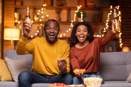 Photo for Emotional beautiful adult black couple spending evening together at home, happy man and woman sitting on couch, drinking beer, eating popcorn and pizza, watching football game, copy space - Royalty Free Image