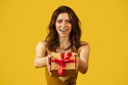 Téléchargez les photos : Holidays celebration. Happy beautiful lady in dress showing wrapped box with gift, posing on yellow background. Young woman feeling happy over special surprise - en image libre de droit