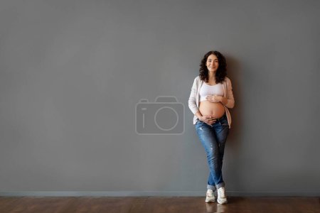 Foto de Pregnancy Ad. Beautiful young pregnant woman embracing belly and smiling at camera, attractive happy expectant lady touching tummy while standing against grey wall indoors, full length shot - Imagen libre de derechos