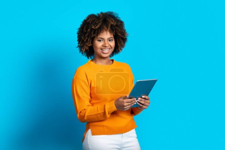 Foto de Happy attractive curly young black woman with teeth braces using modern digital tablet on blue studio background and smiling, checking newest educational or entertaining app, copy space - Imagen libre de derechos