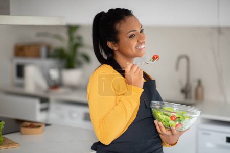 Photo for Cheerful pretty millennial african american lady in apron eat fresh vegetable salad in white kitchen interior, copy space. Weight loss with delicious food, cooking dinner, health care and housekeeping - Royalty Free Image
