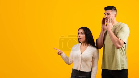Téléchargez les photos : Cheerful shocked young arabic woman and guy in casual point fingers at copy space, isolated on yellow background, studio, panorama. Reaction to good news, surprise, huge sale, emotions, ad and offer - en image libre de droit