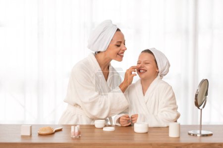 Photo for Happy mother and little daughter in bathrobes and towels applying face cream while having beauty day together at home, young mom playfully touching nose of her preteen female child and laughing - Royalty Free Image
