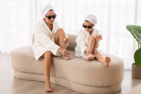 Téléchargez les photos : Happy mother and little daughter in bathrobes and sunglasses making pedicure at home, beautiful young mom and preteen female child applying nail polish on leg fingers and having fun together - en image libre de droit