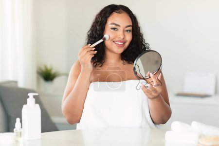 Téléchargez les photos : Makeup, visage concept. Bodypositive lady holding mirror, applying blush and smiling at camera, home interior, copy space. Attractive woman applying nude makeup, using foundation and brush - en image libre de droit