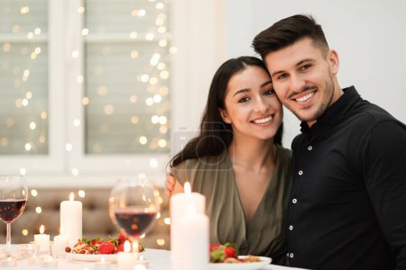 Photo for Portrait of beautiful loving young couple brunette man and woman in nice outwear sit at table decorated with candles, embracing, drinking red wine, celebrating St. Valentines Day at home, copy space - Royalty Free Image