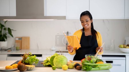 Photo for Smiling pretty millennial black female in apron making salad, reading new recipe in notebook book at table with fresh vegetables in white kitchen interior, panorama. Cooking dinner, household chores - Royalty Free Image