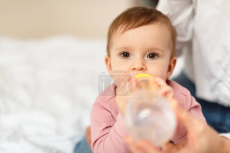 Foto de Closeup shot of cute little infant girl drinking fresh water from bottle, woman holding daughter and helping kid, sitting on bed, free copy space - Imagen libre de derechos