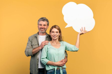 Photo for Excited middle aged woman holding empty speech bubble, having idea, man embracing his wife, posing together over yellow studio background, free space - Royalty Free Image