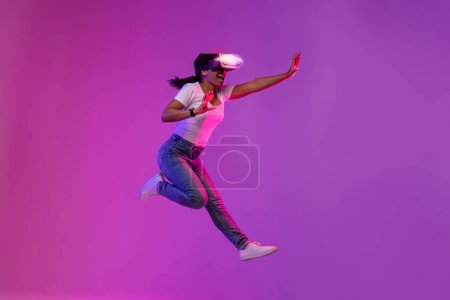 Téléchargez les photos : Excited Young Black Female Wearing Glowing VR Glasses Jumping In Neon Light, Amazed African American Woman Experiencing Virtual Reality And Having Fun Over Purple Studio Background, Copy Space - en image libre de droit