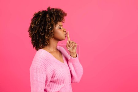 Téléchargez les photos : Serious young black curly female in casual presses her finger to lips and makes shhh sign, isolated on pink background, studio, profile, copy space. Silence, secret gesture, gossip, surprise concept - en image libre de droit