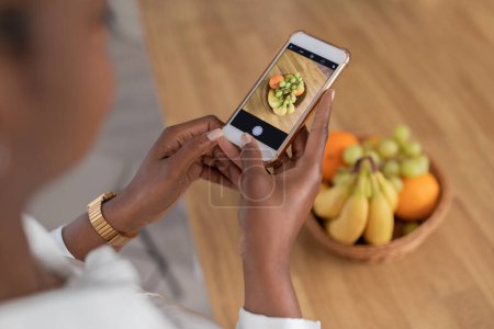 Foto de Cropped of black woman taking photo of bowl of fruits standing on wooden table, using smartphone. Food blogger making content for blog about healthy diet, detox, view above - Imagen libre de derechos