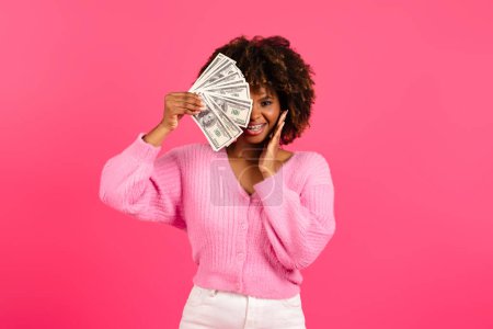 Téléchargez les photos : Smiling pretty young black curly female in casual with braces puts a lot of money to her eye, has fun isolated on pink background, studio, copy space. Cash profit, victory, investment and finance - en image libre de droit