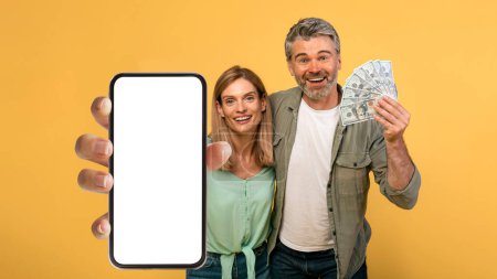 Foto de Giveaway concept. Emotional middle aged couple holding a lot of dollar cash and showing smartphone with blank screen, posing over yellow studio background, mockup - Imagen libre de derechos