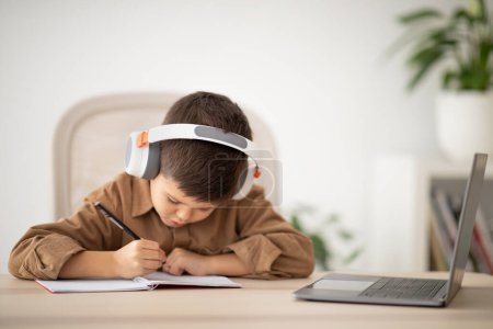 Photo for Busy small european boy in headphones sitting at table, studying, drawing in room interior. Lesson and school, art, learning at home and children garden with device, childhood and education remotely - Royalty Free Image
