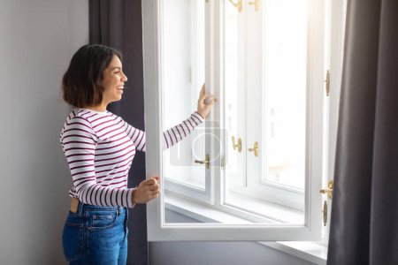 Foto de Portrait Of Young Smiling Arab Woman Opening Window At Home, Happy Millennial Middle Eastern Female Unlocking Frame And Looking Out, Enjoying View And Breathing Fresh Air, Copy Space - Imagen libre de derechos