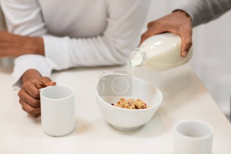Téléchargez les photos : Cropped of black couple in pajamas have breakfast together at home. Unrecognizable african woman holding mug with tea or coffee, man pouring milk into granola bowl. Diet, nutrition concept - en image libre de droit