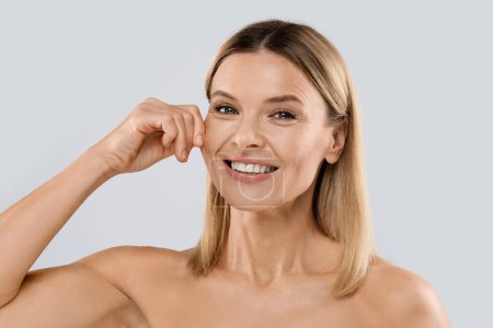 Téléchargez les photos : Anti-aging treatment in cosmetology. Smiling attractive half-naked middle aged blonde woman touching her cheeks, demonstrating youthful smooth elastic skin, posing on grey studio background, closeup - en image libre de droit