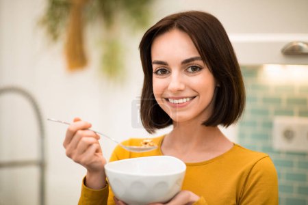 Foto de Closeup shot of cheerful smiling pretty brunette young woman enjoying healthy breakfast at home, eating cereals, whole grain granola, oatmeal, copy space. Nutrition, diet concept - Imagen libre de derechos