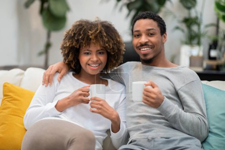Téléchargez les photos : Cute positive beautiful african american couple in homewear sitting together on couch in living room, drinking coffee, embracing and smiling at camera, enjoying lazy weekend at home - en image libre de droit