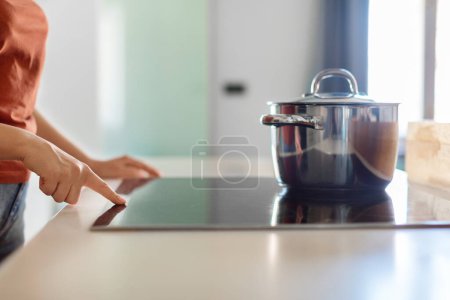 Foto de Female hand turning on induction stove with steel cooking pan in kitchen, closeup shot of woman finger touching sensor button on electrical hob while preparing food at home, side view, cropped - Imagen libre de derechos