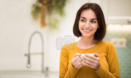 Téléchargez les photos : Pretty cheerful smiling brunette young woman holding cup of tea, looking at copy space, cozy modern kitchen interior. Beautiful lady enjoying morning coffee at home, panorama - en image libre de droit