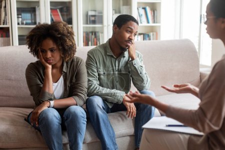 Foto de Frustrated married young black couple attending family therapist, man and woman sitting on couch back to back, avoid looking at each other while listening to female psychologist - Imagen libre de derechos