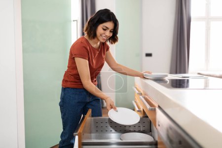 Téléchargez les photos : Beautiful young arab woman tidying up kitchen cupboard during general cleaning, happy middle eastern housewife neatly placing clean dishes in drawer after washing, enjoying domestic chores - en image libre de droit