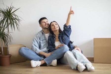 Photo for Happy Young Couple Sitting On Floor Among Cardboard Boxes After Moving And Pointing Up, Cheerful Spouses Imagining Home Interior Design And Discussing Decoration After Relocation To Own House - Royalty Free Image