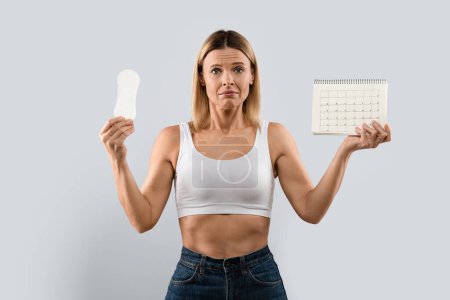 Foto de Unhappy depressed middle aged woman being in bad mood, suffers from premenstrual syndrom, awaits for critical days soon, demonstrates periods calendar and sanitary napkin. Women healthcare concept - Imagen libre de derechos