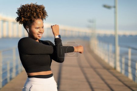 Téléchargez les photos : Outdoor Workout. Sporty Black Woman Training Outside On Wooden Pier, Stretching Arm Muscles, Athletic African Female Wearing Smartwatch And Wireless Easphones Warming Up Before Jogging, Copy Space - en image libre de droit