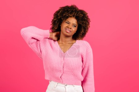 Téléchargez les photos : Despaired sad tired young black curly female in casual suffering from neck pain, make massage, isolated on pink background. Health problems, muscle pain after training, overwork and arthritis, injury - en image libre de droit