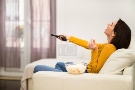Foto de Happy attractive young brunette woman in casual outfit sitting on couch in living room, holding remote, eating popcorn and laughing, enjoying TV show, home interior, side view, copy space - Imagen libre de derechos