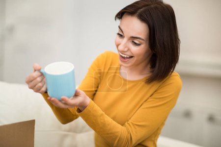 Foto de Excited surprised happy pretty brunette young woman in casual outfit sitting on couch, holding blue china mug, checking birthday or xmas presents, home interior, copy space - Imagen libre de derechos