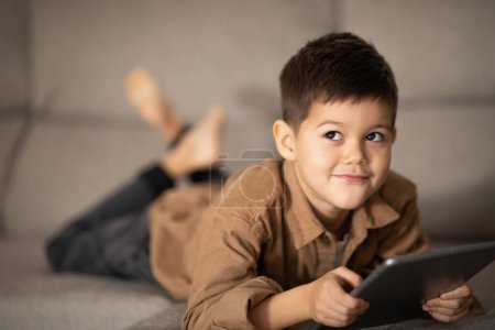 Téléchargez les photos : Pensive cheerful small european boy lies on sofa, thinks with tablet, look at free space in room interior. Study at home with device, childhood and upbringing remotely, watch videos and play games - en image libre de droit