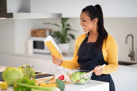 Photo for Cheerful millennial african american woman in apron reads new recipe in notebook or book, make salad at table with organic fresh vegetables in white kitchen interior. Cooking homemade dinner at home - Royalty Free Image