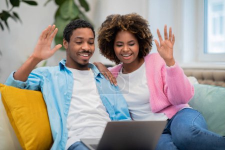 Photo for Positive cheerful millennial black spouses attractive man and woman in casual sitting on couch, waving at laptop screen, have online party with friends while staying home, copy space - Royalty Free Image