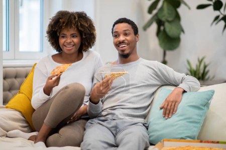Photo for Happy beautiful millennial black lovers sitting on couch, eating pizza, looking aside and smiling, loving african american man and woman spending weekend at home, have party, copy space - Royalty Free Image